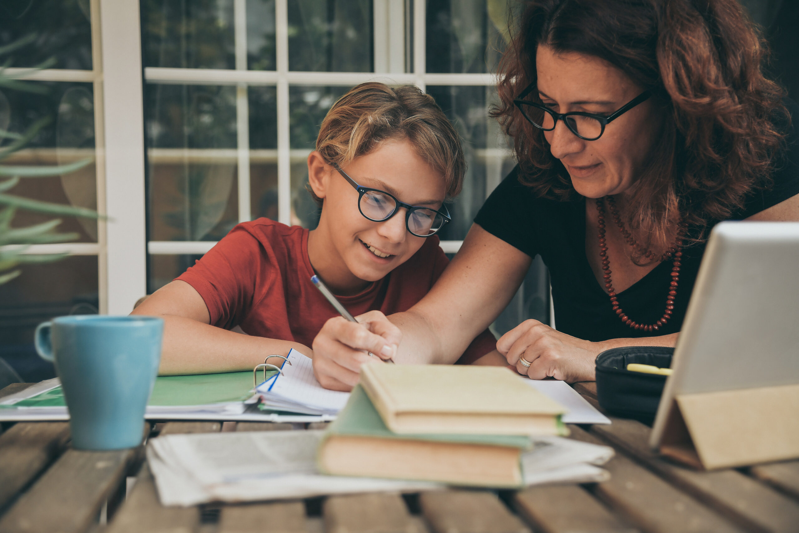 Young student doing homework at home with school books, newspaper and digital pad helped by his mother. Mum writing on the copybook teaching his son. Education, family lifestyle, homeschooling concept