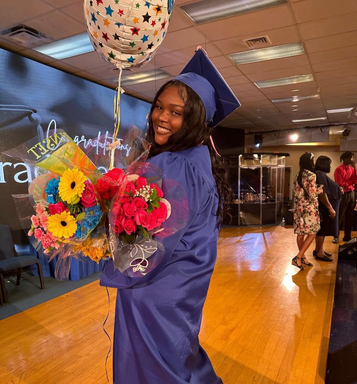 Picture of an African American girl in her cap and gown. She's holding flowers and a balloon. She's looking back towards the camera.